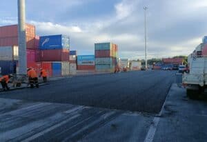 Construction workers and machinery operate on a freshly paved surface at a shipping yard surrounded by large stacks of colourful shipping containers. The scene takes place under a partly cloudy blue sky, showing an active industrial environment.
