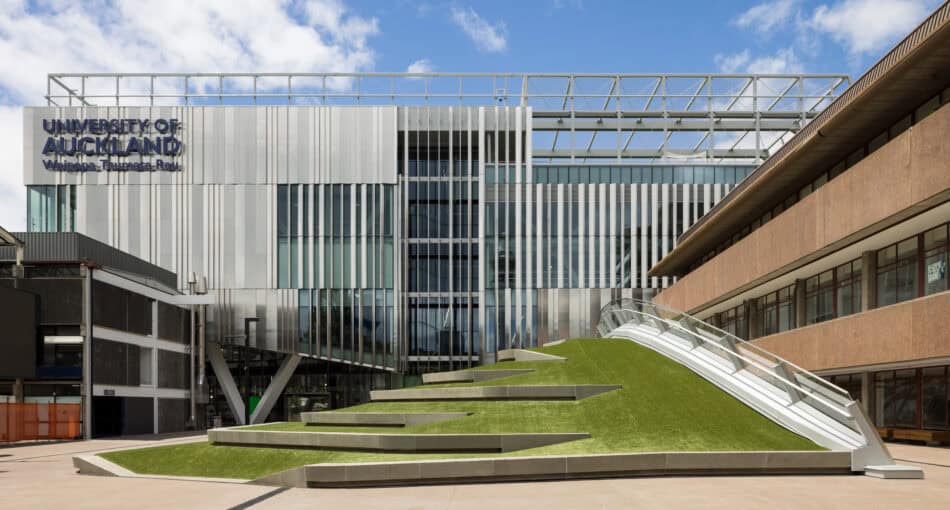 Modern building at the University of Auckland’s Waipapa Taumata Rau campus featuring a sleek façade of vertical metal fins and glass panels. In the foreground is a distinctive green, stepped grass mound with a curved ramp and handrail leading up to the building. The sky is clear with scattered clouds.