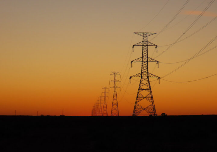 A row of high-voltage power transmission towers stretches into the distance across a flat landscape at sunset. The sky is a gradient of warm orange and soft grey tones, creating a striking silhouette of the towers and power lines against the fading light.