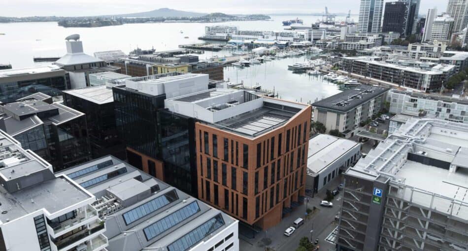 Aerial view of Auckland’s Wynyard Quarter waterfront precinct featuring modern commercial buildings and marinas. In the centre is a prominent mid-rise building with a red brick façade and vertical window patterns, surrounded by glass and metal structures. The harbour and boats are visible in the background under an overcast sky.