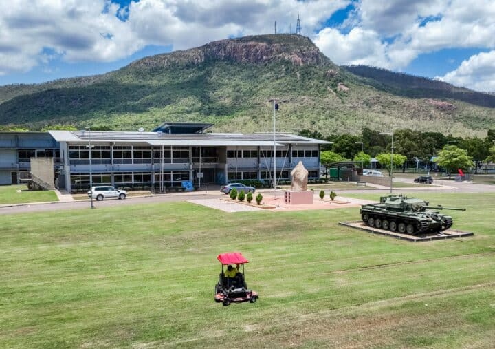 A ride-on mower trims the large lawn outside a Defence facility, where a tank monument and a flagpole stand near the main building. Behind the site, forested mountains rise under a partly cloudy sky.