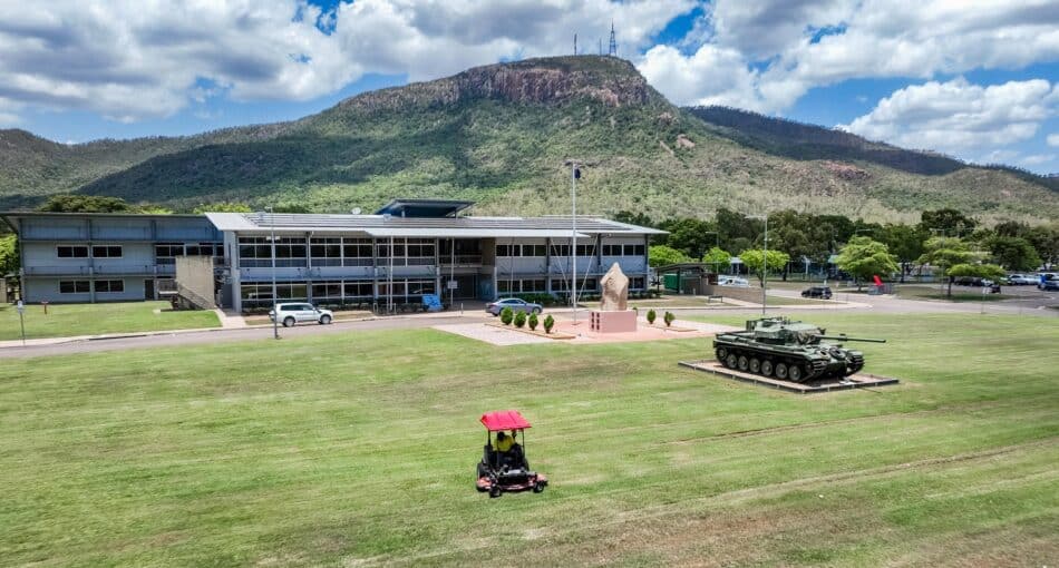 A ride-on mower trims the large lawn outside a Defence facility, where a tank monument and a flagpole stand near the main building. Behind the site, forested mountains rise under a partly cloudy sky.