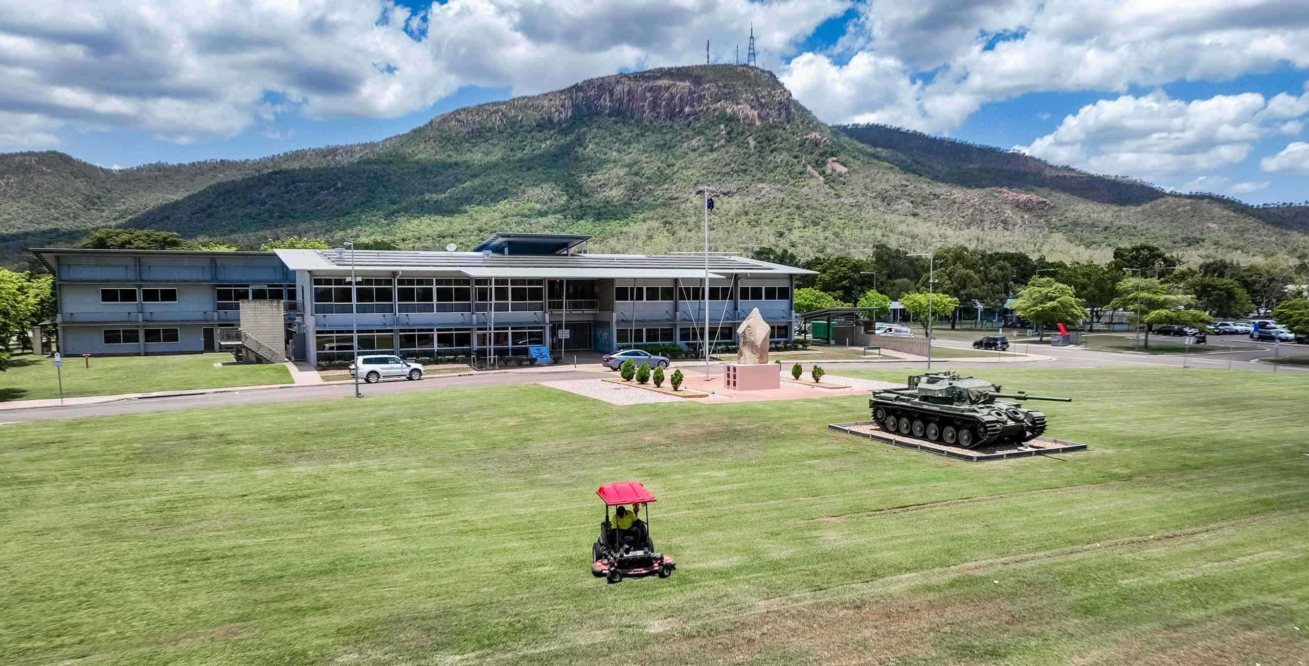 A ride-on mower trims the large lawn outside a Defence facility, where a tank monument and a flagpole stand near the main building. Behind the site, forested mountains rise under a partly cloudy sky.